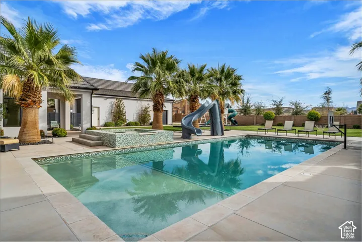 In-ground swimming pool featuring a built-in slide, raised spa with mosaic tile finish, expansive concrete patio, and mature palm trees