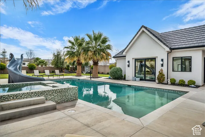 Backyard featuring a swimming pool with a mosaic tile-finished spa, a slide, concrete decking, and mature palm trees
