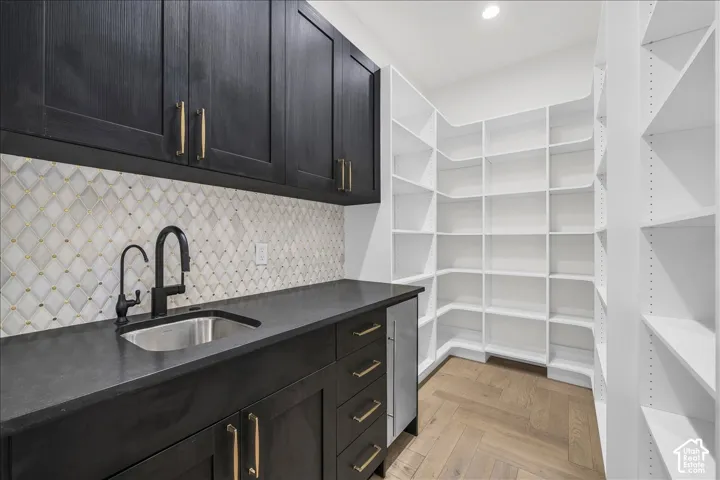 Walk-in pantry featuring white floor-to-ceiling shelving, dark cabinetry with brass-finish hardware, a matte black faucet, a stainless steel sink, and wood-finish flooring in a herringbone pattern