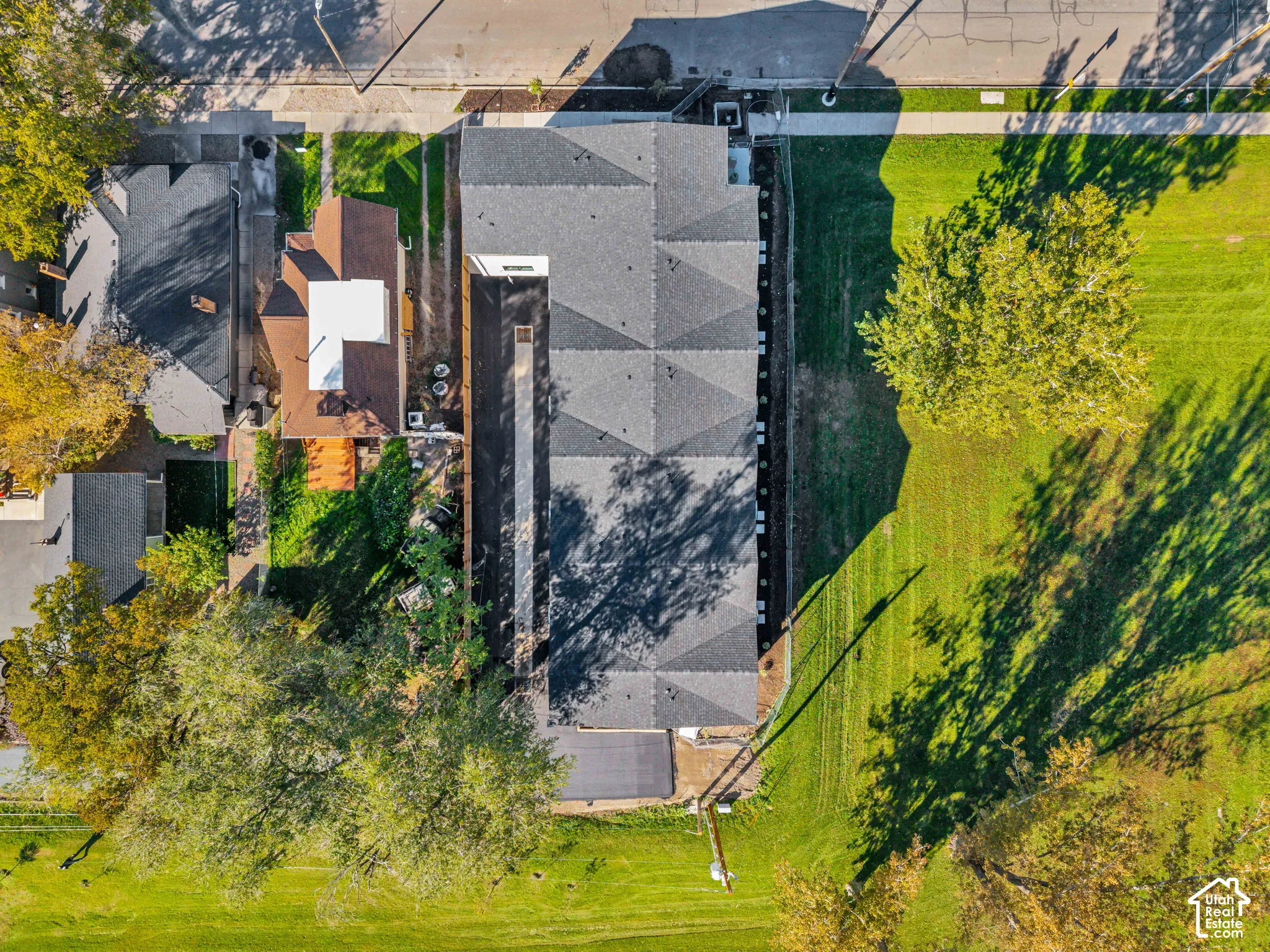 Aerial perspective of the property featuring a multi-sectioned asphalt shingle roof, concrete driveway, and expansive lawn with mature trees