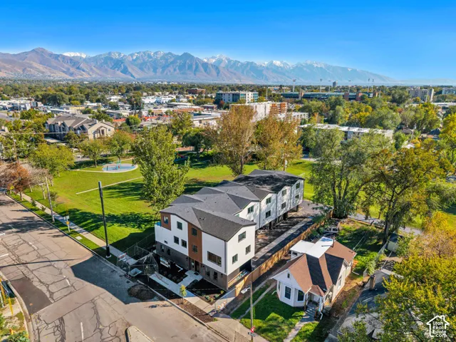 Aerial perspective featuring a multi-level structure with a dark shingle roof, wood-tone paneling, and stucco exterior, situated near an open green space with mature trees against a mountain backdrop