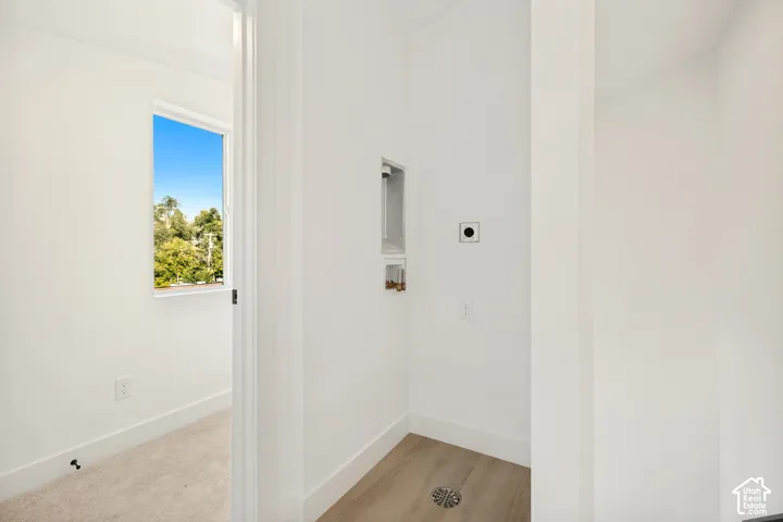 Utility area featuring wood-finish flooring, a recessed water supply box, a drain, and a dedicated electrical outlet