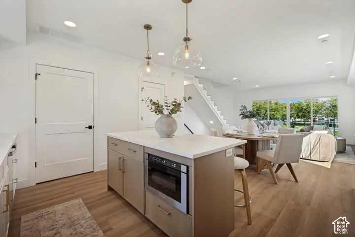 Kitchen island featuring a stone-composite countertop, light wood cabinetry, and a built-in stainless steel microwave, complemented by dual glass globe pendant lights and wide-plank wood-finish flooring