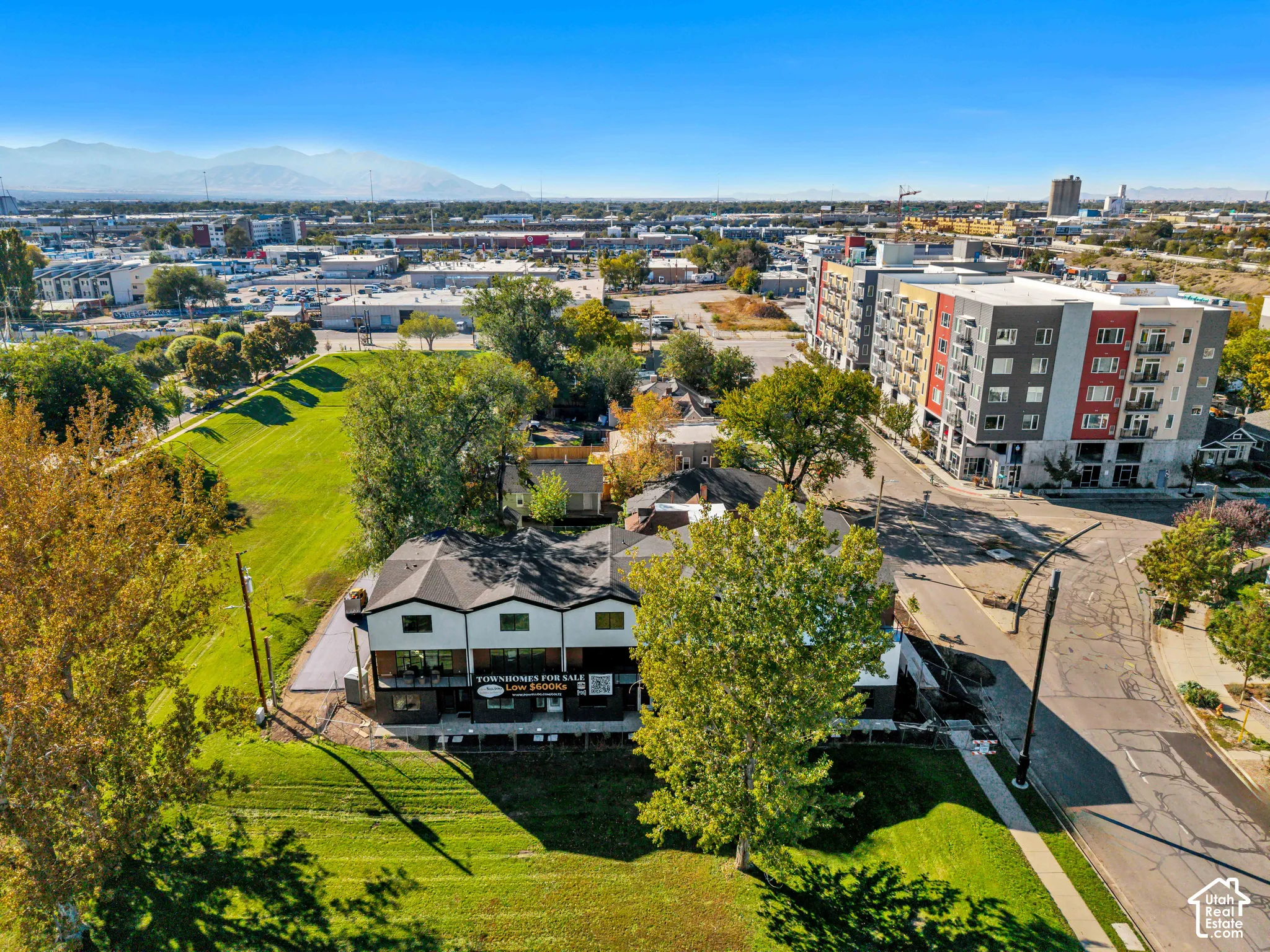 Aerial perspective of multi-level townhome featuring a dark shingle roof, contrasting light and dark exterior siding, ground-level paved patio, and mountain backdrop