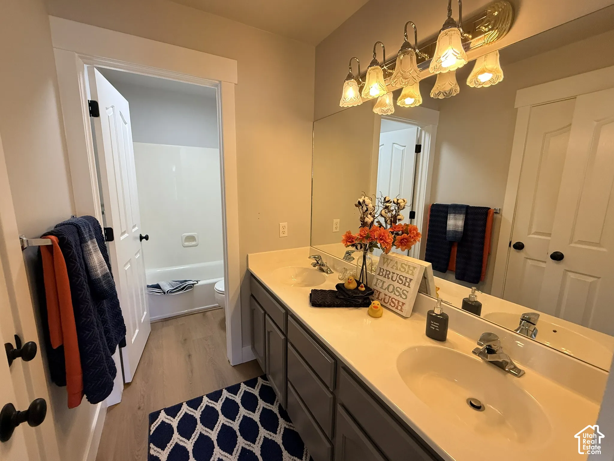 Dual-sink vanity with light-toned countertop, custom cabinetry, wall-to-wall vanity mirror, multi-light fixture, and wood-finish flooring