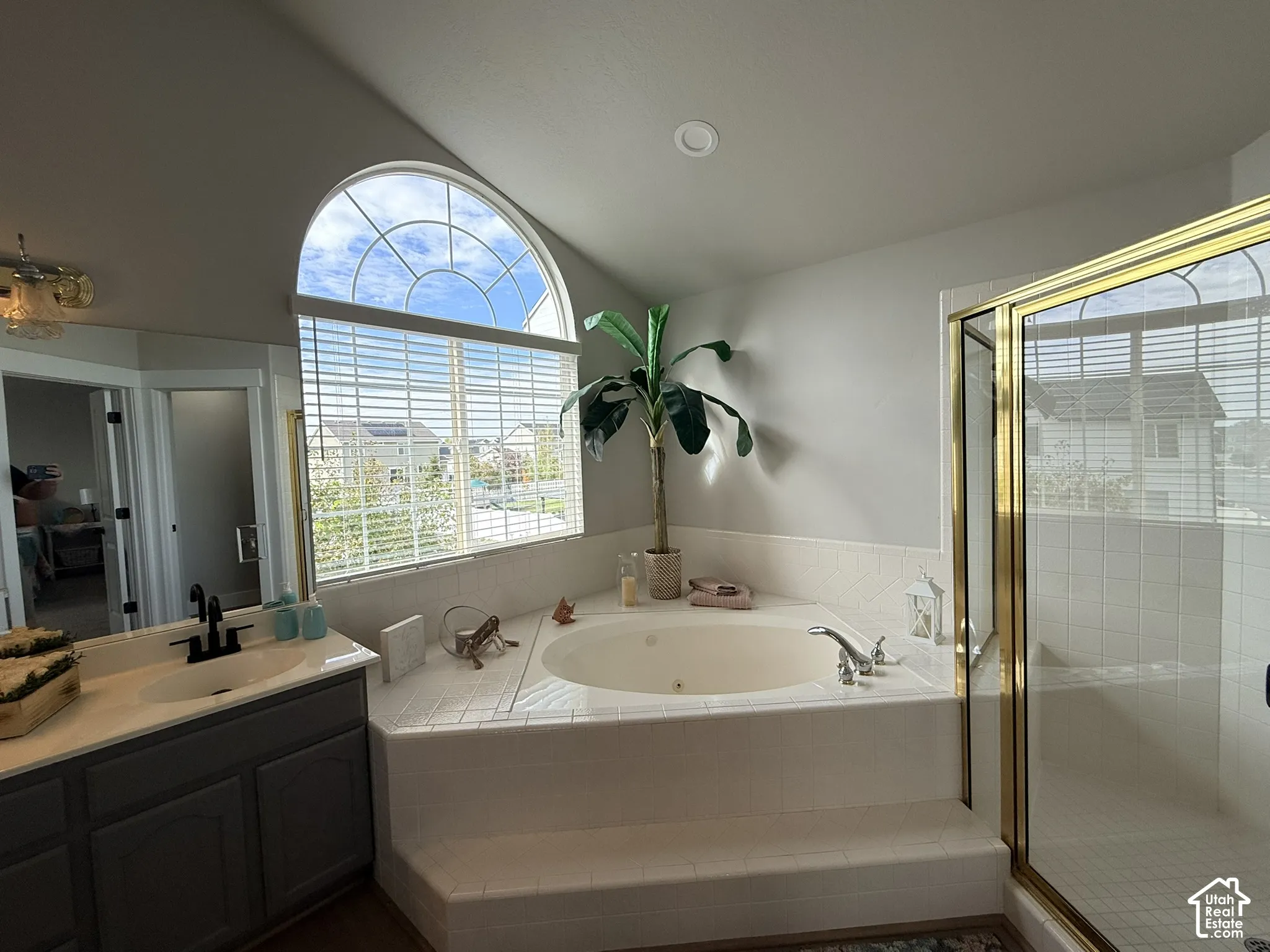 Arched window, corner jetted soaking tub with tile surround, glass-enclosed shower with gold-tone frame, and grey vanity cabinetry featuring a light-toned countertop with a matte black faucet