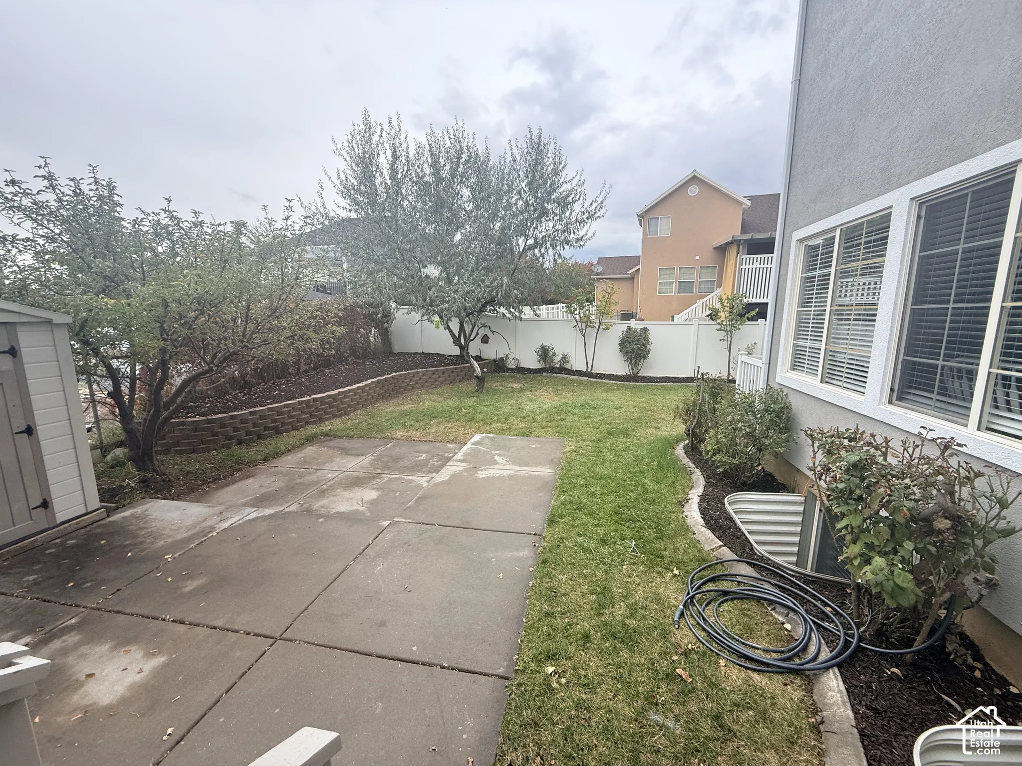 Backyard featuring a concrete patio, stone-block retaining wall, lawn, mature trees, and white vinyl perimeter fencing