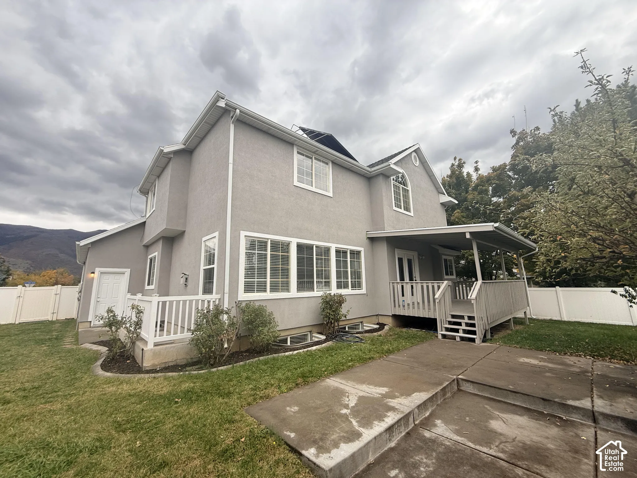 Two-story exterior with light gray stucco siding, covered wood deck, concrete patio, solar panels, and white vinyl privacy fencing