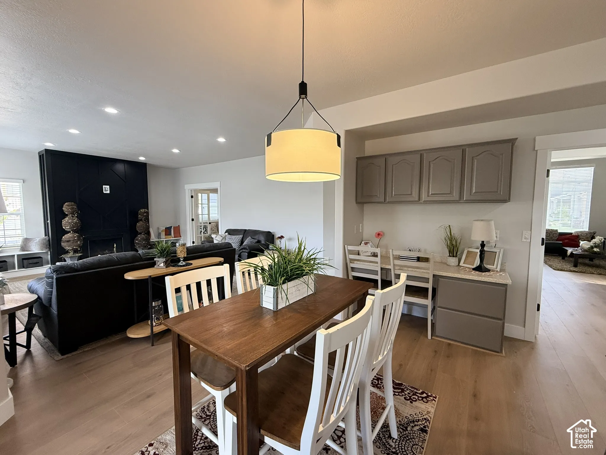 Open-concept area featuring wood-finish flooring, a contemporary drum pendant light, a built-in workstation with neutral-toned cabinetry, and a floor-to-ceiling dark-toned fireplace surround