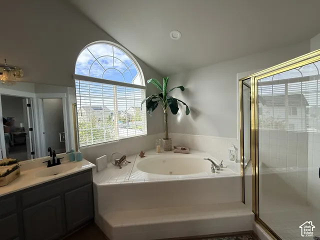 Arched window, corner jetted soaking tub with tile surround, glass-enclosed shower with gold-tone frame, and grey vanity cabinetry featuring a light-toned countertop with a matte black faucet