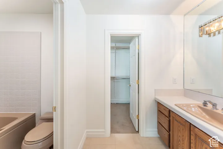 Bathroom featuring a built-in wood vanity with a solid-surface countertop and integrated sink, a full-wall vanity mirror with multi-bulb light fixture, a tiled tub surround, and a walk-in closet with built-in shelving