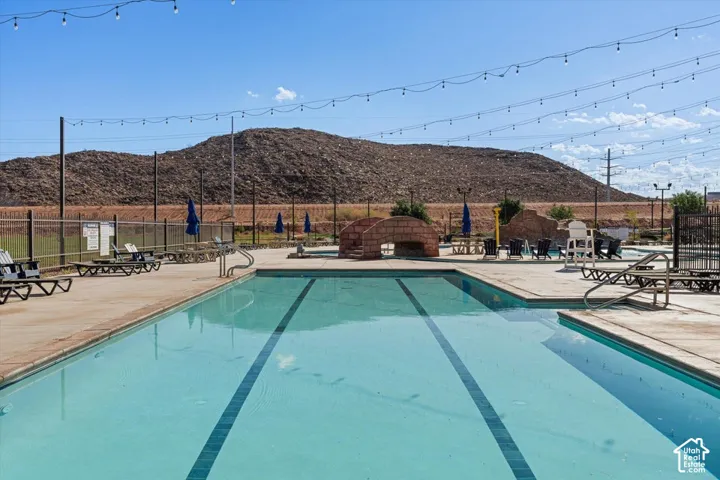Swimming pool with lane markings, stone-textured footbridge, concrete decking, metal perimeter fencing, and backdrop of mountain terrain