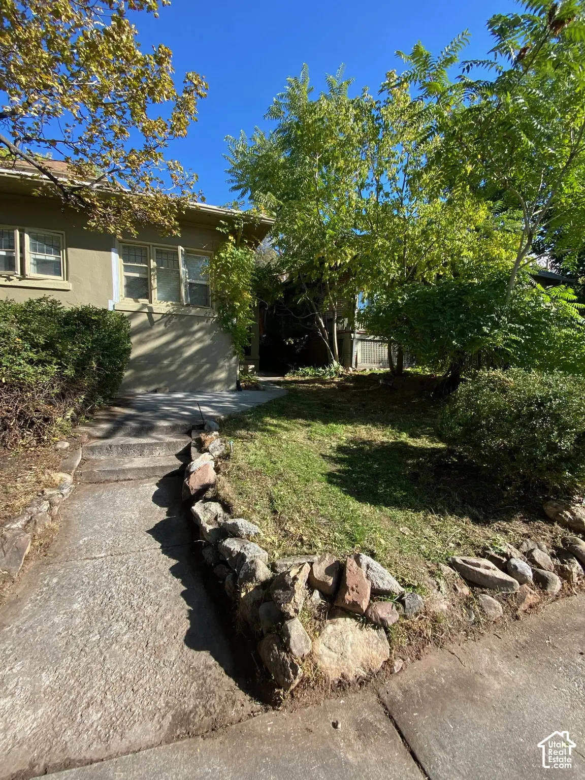 Exterior elevation featuring stucco siding, multi-pane windows, a stone-lined walkway, and natural stone landscaping elements
