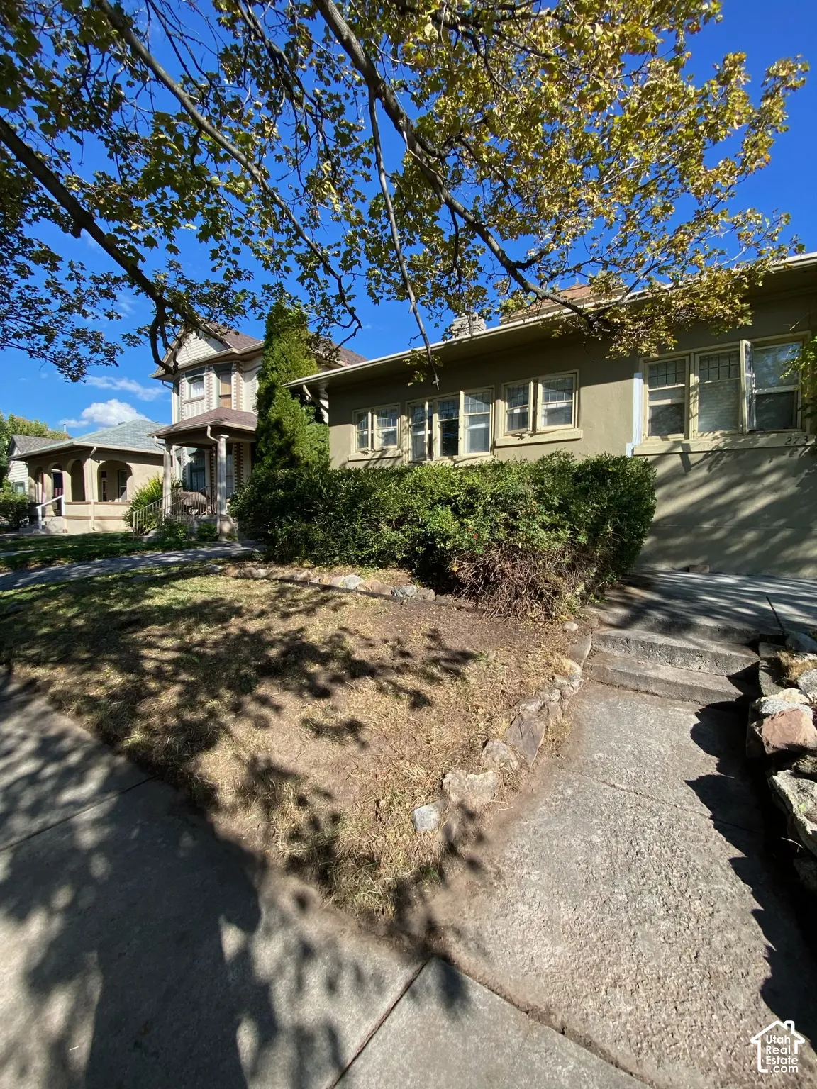 Stucco exterior featuring multi-pane windows, mature trees, stone retaining wall, and concrete walkway