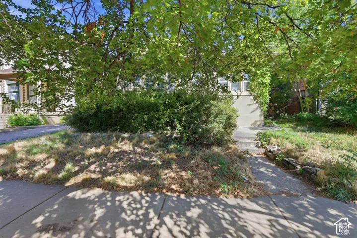 Exterior facade featuring a concrete walkway, mature trees, established shrubs, and stone retaining wall elements