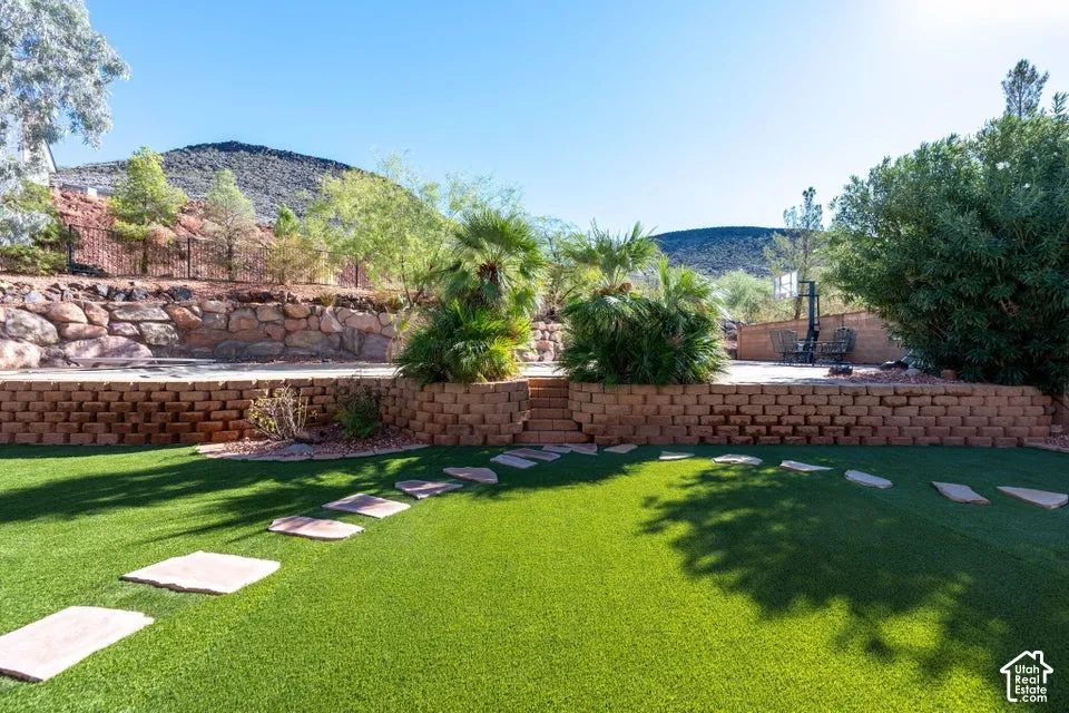 Terraced backyard featuring synthetic turf, stone-paver walkway, stacked-stone retaining walls, mature vegetation, and a mountain backdrop