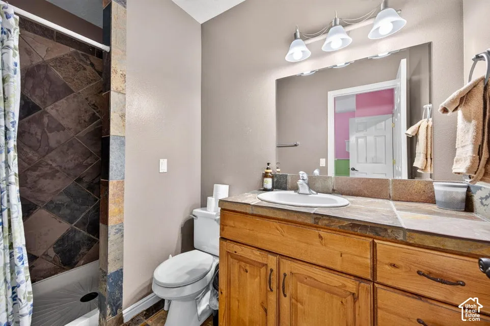 Bathroom featuring a wood vanity with a stone-tile countertop, integrated basin, three-light fixture, and a walk-in shower with decorative tile surround