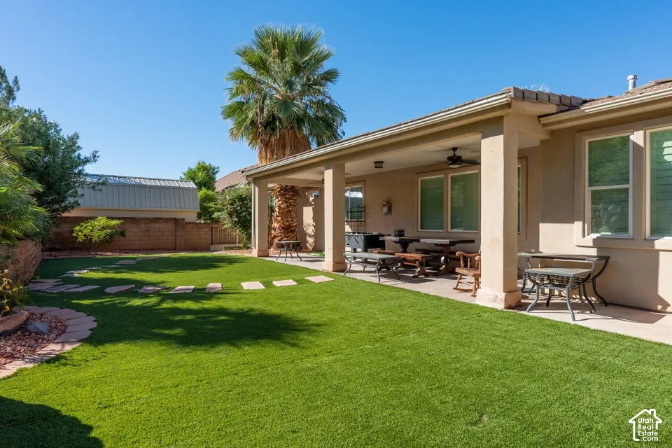 Covered concrete patio with a ceiling fan, synthetic turf lawn, stone paver pathway, stucco exterior, and masonry perimeter wall