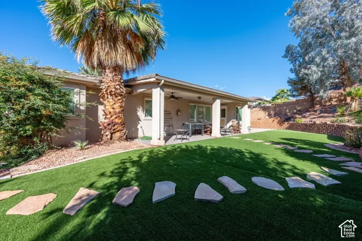 Covered concrete patio with a ceiling fan, synthetic turf lawn, stone paver pathway, and tiered masonry retaining wall
