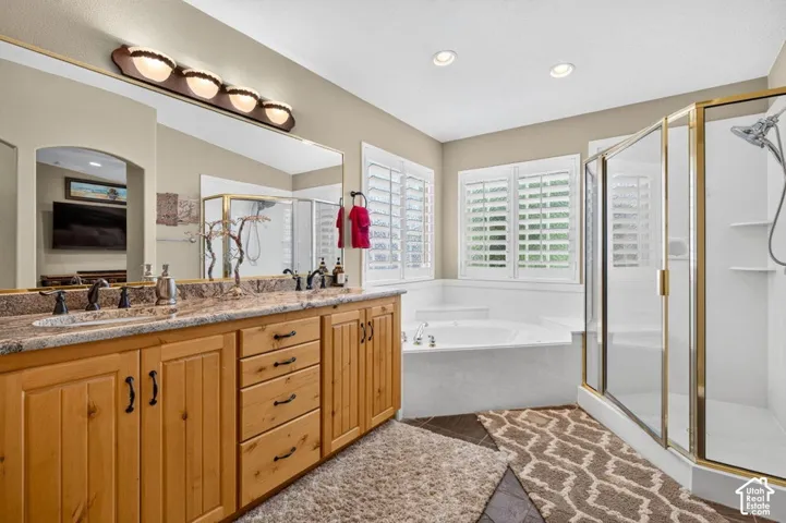 Bathroom featuring a dual-sink vanity with stone-surface countertops, recessed lighting, plantation shutters, a corner soaking tub, and a glass-enclosed shower
