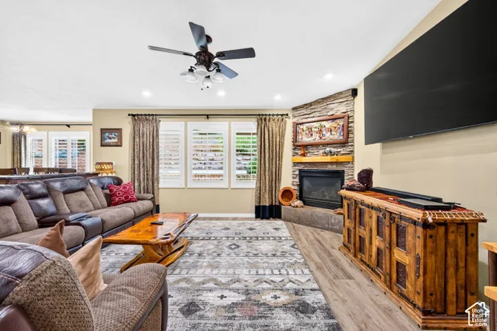 Corner fireplace with stone-veneer surround and timber mantel, wood-finish flooring, plantation shutters, and a ceiling fan with integrated lighting