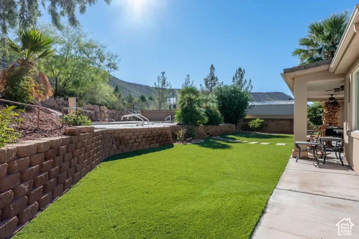 Backyard featuring an inground swimming pool, concrete covered patio, synthetic turf lawn, stacked stone retaining wall, and mountain backdrop