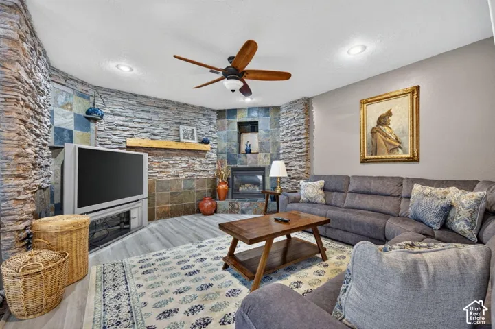 Textured stone accent wall featuring a corner fireplace with square tile surround, wood-finish flooring, recessed lighting, and a ceiling fan