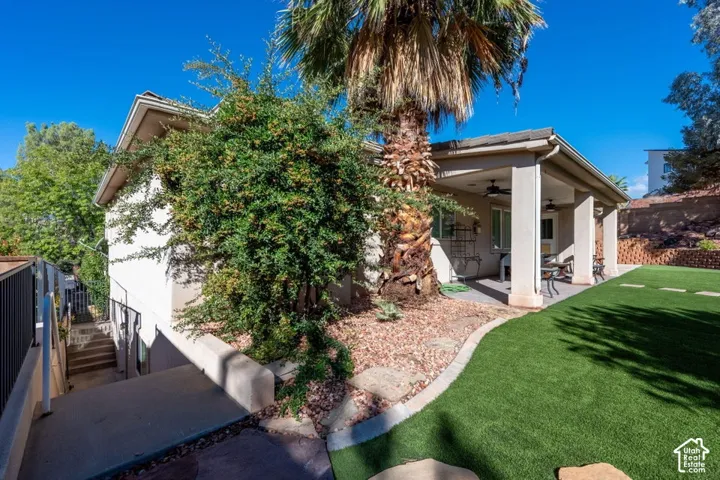 Covered rear patio with ceiling fans, artificial turf lawn, concrete walkways, and tiered landscaping