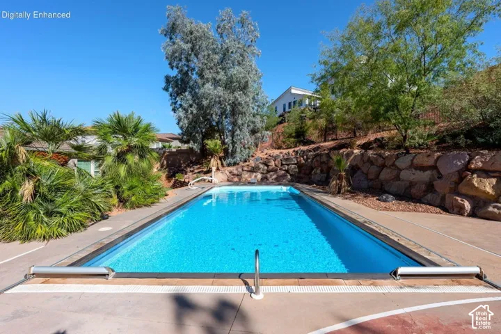 Rectangular swimming pool featuring a concrete deck, built-in pool cover, and stone retaining wall with mature landscaping