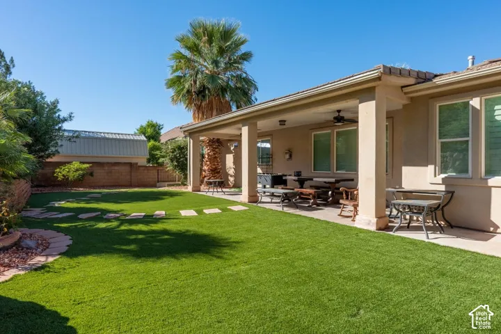 Covered concrete patio with a ceiling fan, synthetic turf lawn, stone paver pathway, stucco exterior, and masonry perimeter wall
