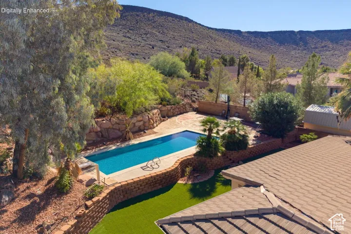 Backyard featuring a rectangular swimming pool, tiered stone retaining walls, artificial turf lawn, concrete patio, and mature trees with a mountain backdrop