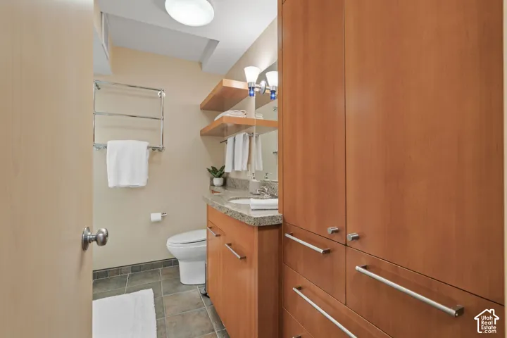 Bathroom featuring wood-finish cabinetry, stone-surface countertop, integrated wall-mounted shelving, dual-light vanity fixture, and tiled flooring