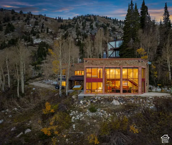 Multi-level mountain residence featuring expansive floor-to-ceiling windows, natural stone exterior cladding, and steel structural framing, set against a backdrop of mountainous terrain and mature aspen trees