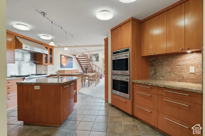 Kitchen featuring warm-toned wood cabinetry, stone countertops, stainless steel double wall ovens, a matching range hood, and tile flooring