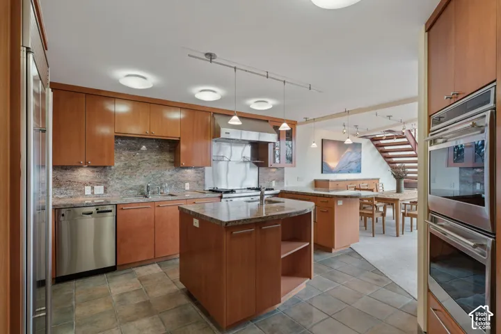 Kitchen featuring cabinetry with a natural wood finish, stone countertops, stainless steel double wall ovens, a stainless steel gas range with a metal backsplash, and a central island with an integrated sink