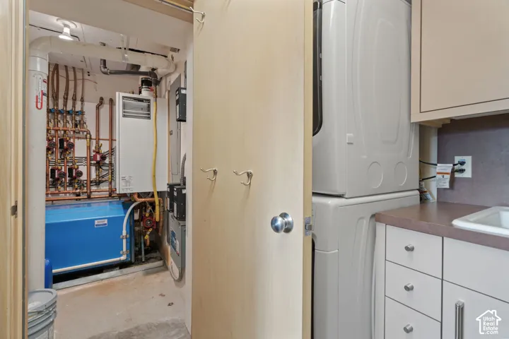 Utility space featuring a stacked laundry unit, white cabinetry with metallic hardware, integrated sink, and mechanical systems including a boiler and copper plumbing components