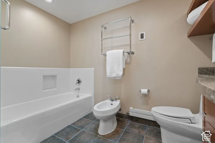 Bathroom featuring stone-finish tile flooring, a bathtub with a recessed wall niche, a bidet, a wall-mounted chrome towel warmer, and a vanity with a solid-surface countertop