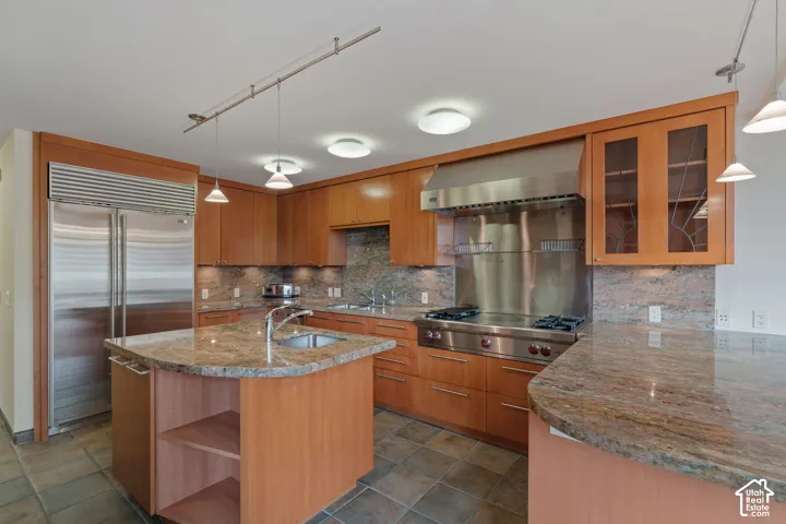 Kitchen featuring wood cabinetry, stone countertops, integrated stainless steel refrigerator, professional-grade gas range with stainless steel backsplash, and tile flooring
