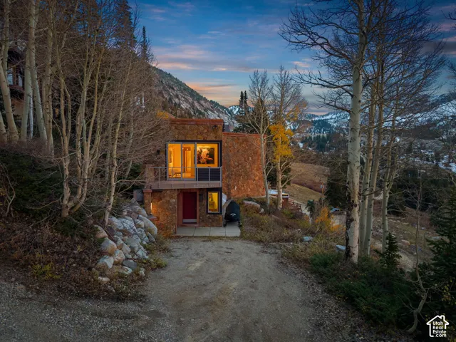 Mountain residence featuring extensive natural stone exterior cladding, expansive floor-to-ceiling glass windows, an elevated metal-railing balcony, and a paved stone entry path nestled within a wooded landscape
