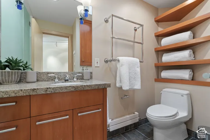 Vanity featuring a stone-composite countertop, undermount sink, and wood-finish cabinetry, paired with wall-mounted shelving, a metal towel warmer, and dark tile flooring