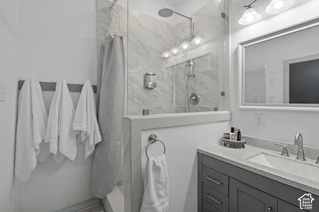 Glass-enclosed shower with a rainfall showerhead, tiled shower surround, framed mirror, vanity with a light-colored countertop, and a wall-mounted light fixture