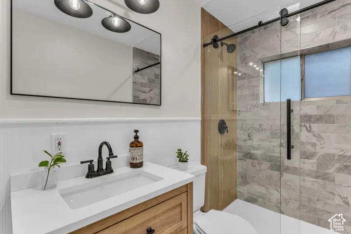 Bathroom featuring a wood-finish vanity with a white countertop, walk-in shower with stone-patterned tile, glass sliding enclosure, and matte black fixtures