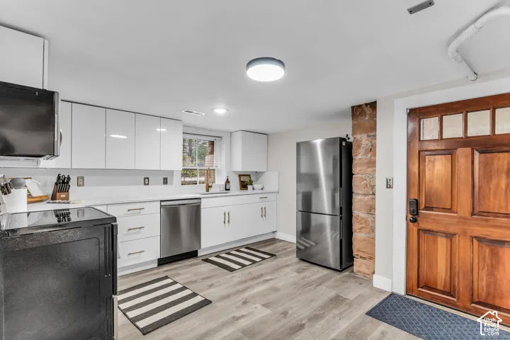 Kitchen featuring white flat-panel cabinetry, light-toned wood-finish flooring, stainless steel appliances, exposed stone pillar detail, and a six-panel wood entry door with glass accents