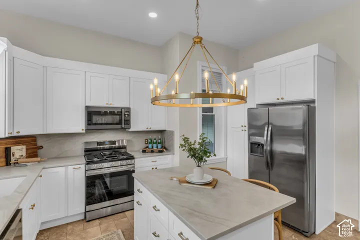 Kitchen featuring white shaker-style cabinetry, stone-finish countertops, stainless steel appliances, a central island, and a circular brass chandelier