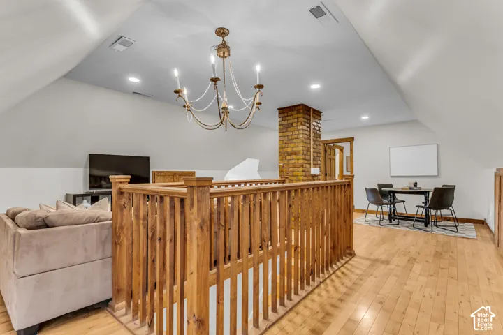 Wood-finish flooring, vaulted ceilings, exposed brick chimney, and a multi-tiered candelabra chandelier anchored by a wood railing
