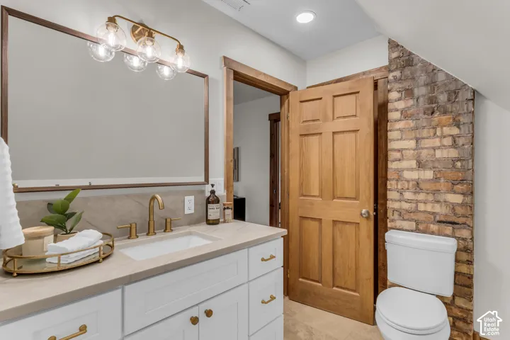 Bathroom vanity featuring white shaker-style cabinetry, light-toned countertop with an integrated rectangular sink, brass hardware, wood-framed mirror, and an exposed brick wall accent