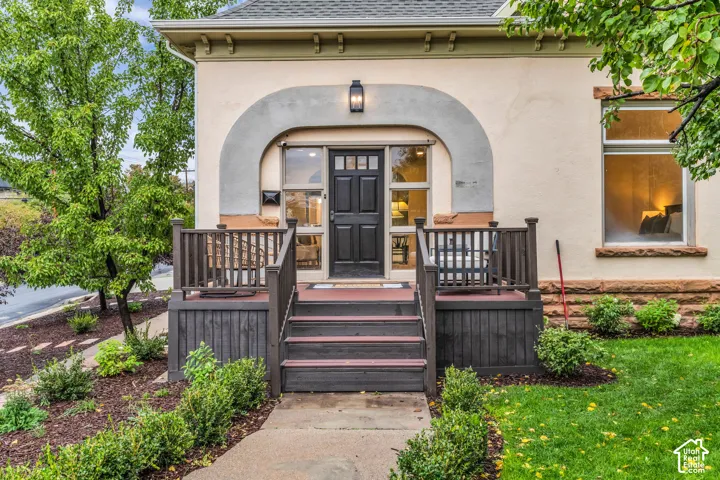 Stucco exterior featuring an arched entryway, raised wood-finish deck with railings, stone foundation detailing, and multi-light entrance door