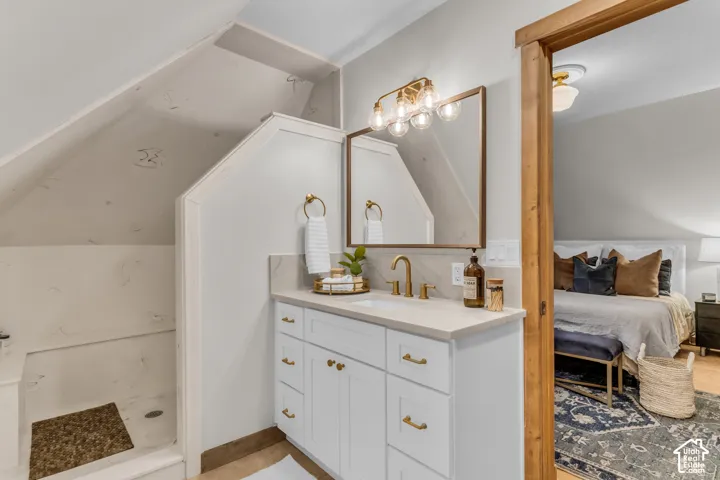 White vanity with brass hardware, neutral countertop, rectangular undermount sink, multi-light fixture, and walk-in shower with stone-style wall surround