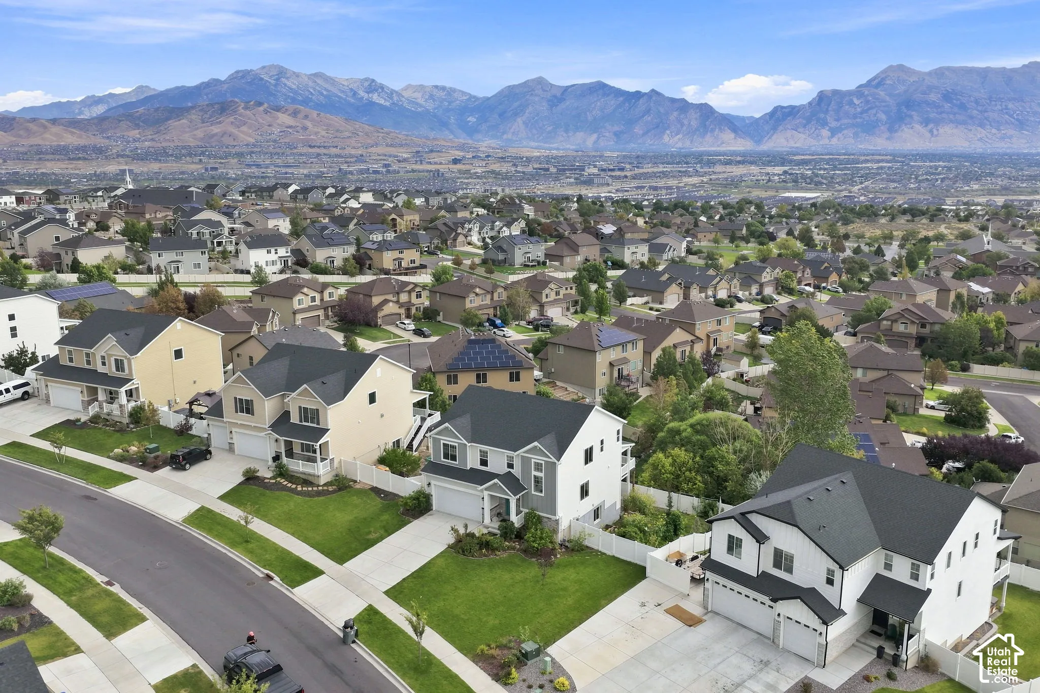 Aerial perspective featuring residential lots with manicured lawns, paved driveways, multi-story single-family homes with asphalt shingle roofs, and a backdrop of mountain ranges
