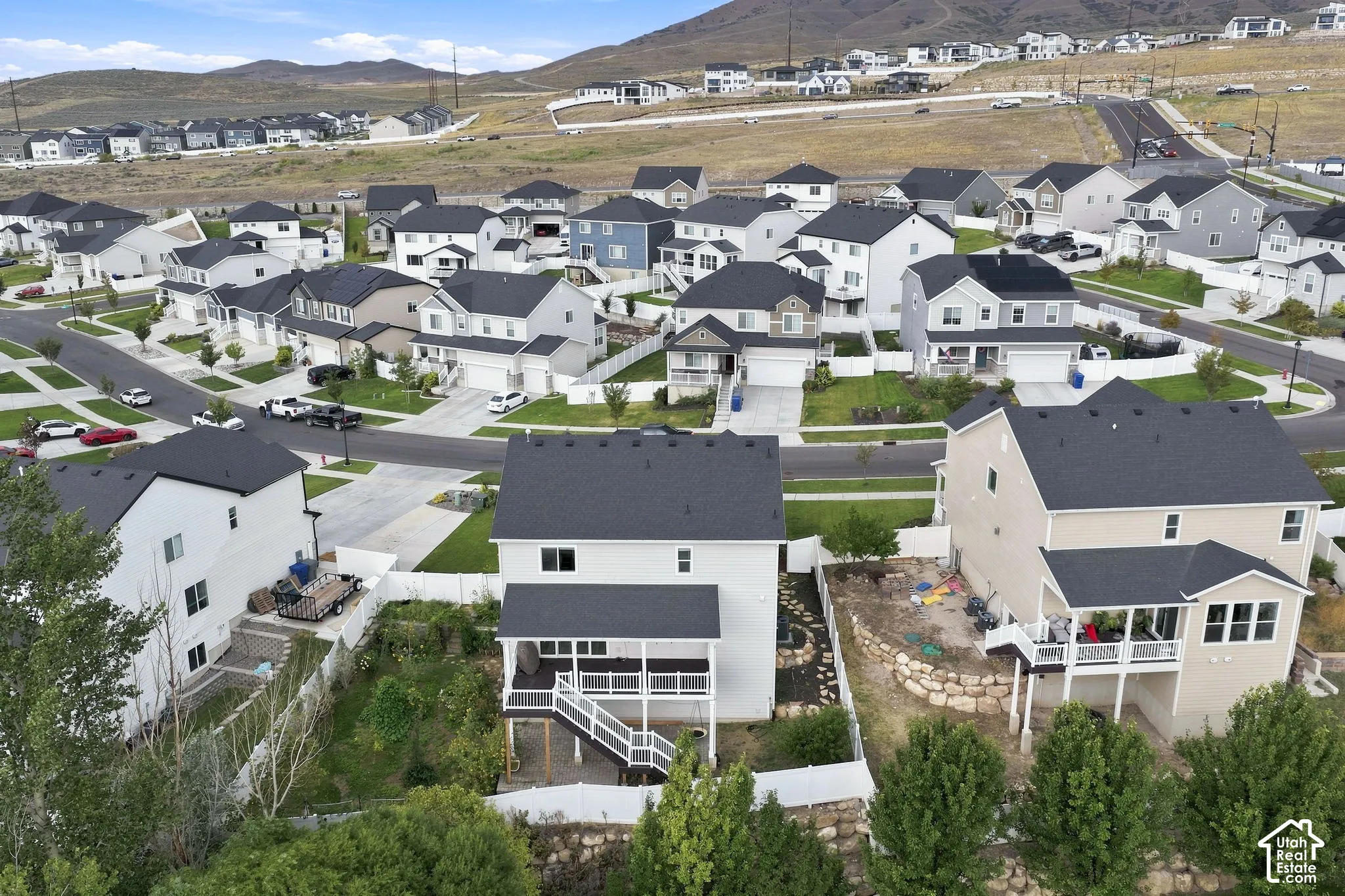 Aerial perspective of a multi-level suburban residence featuring a raised wooden deck with a staircase, stone retaining wall, white vinyl fencing, and mountain terrain backdrop